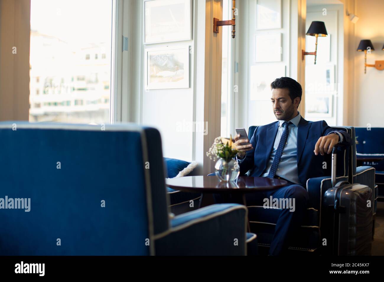 Uomo d'affari seduto nella lobby dell'hotel con valigia, usando il telefono cellulare. Foto Stock