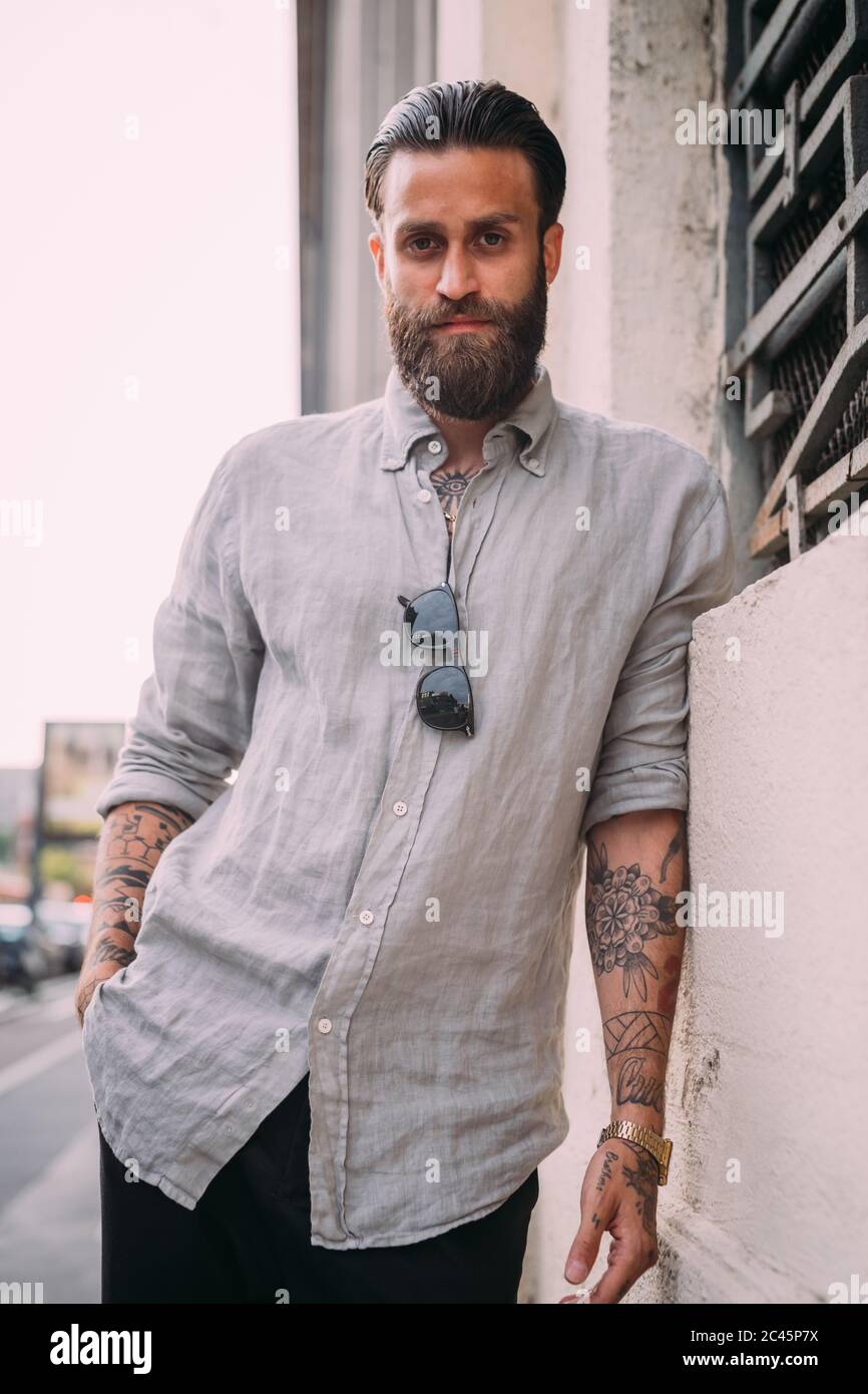 Ritratto di giovane bearded con capelli marroni, con tatuaggi sulle braccia, con camicia grigia, appoggiato a parete. Foto Stock