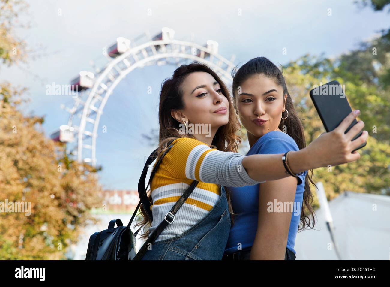 Due giovani donne con lunghi capelli marroni in piedi in un parco vicino a una ruota panoramica, prendendo selfie con il telefono cellulare. Foto Stock
