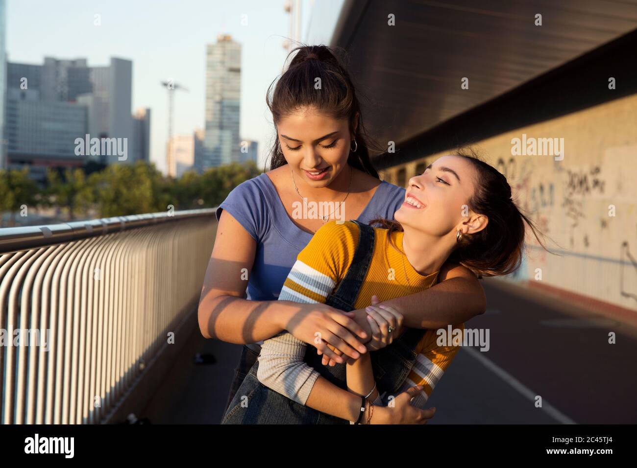 Due giovani donne con lunghi capelli marroni in piedi sul ponte urbano, abbraccianti e sorridenti. Foto Stock