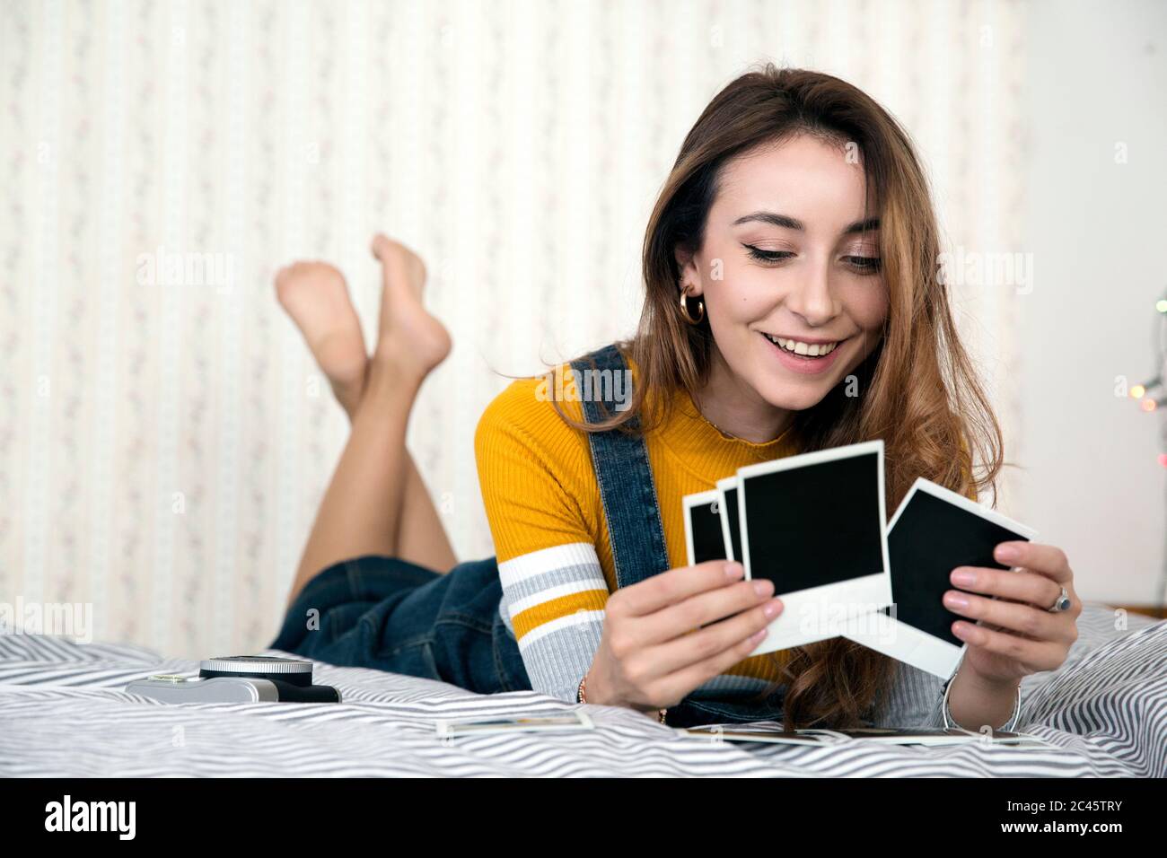 Giovane donna con lunghi capelli marroni sdraiata sul letto, guardando le fotografie Polaroid. Foto Stock