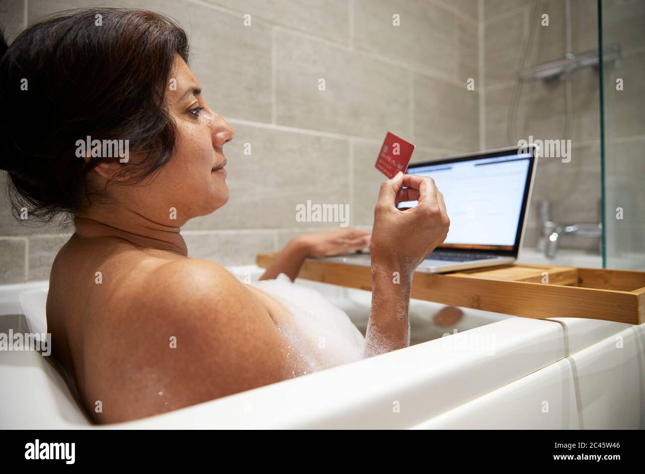 Donna seduta nella vasca da bagno, avendo bagno di schiuma e shopping online sul suo laptop durante la crisi di Coronavirus. Foto Stock