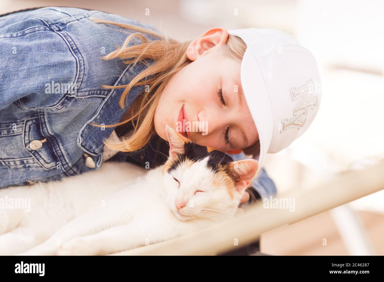 Giovane bionda, età della scuola primaria, indossare un cappello da sole, carezzare il gatto sdraiato sul lettino Foto Stock