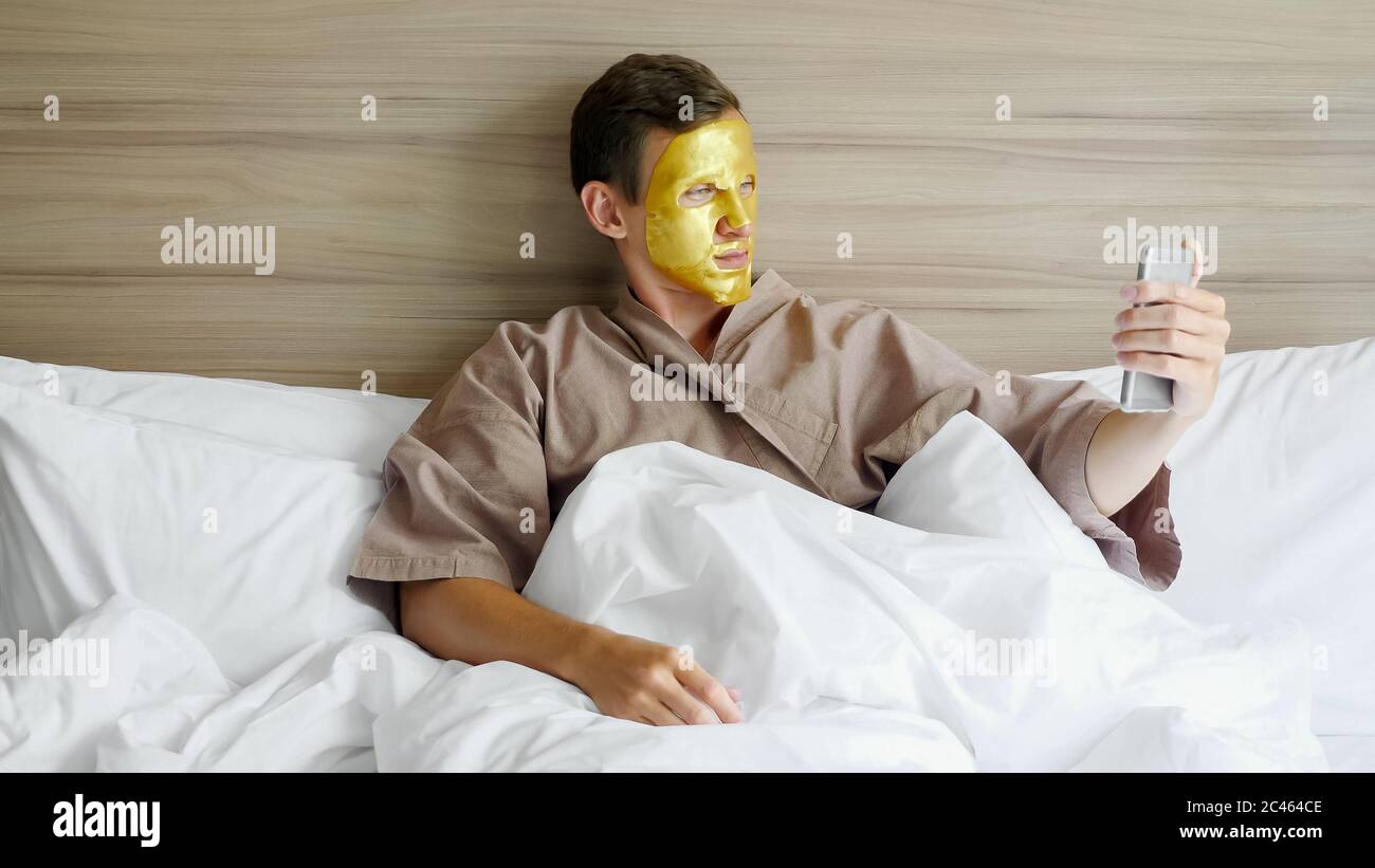 divertente ragazzo annoiato in cappotto marrone con maschera dorata faccia  rende selfie sdraiato in morbido letto in camera d'albergo Foto stock -  Alamy