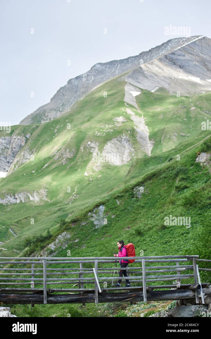 Vista laterale del viaggiatore signora con zaino che cammina sul ponte con colline erbose sullo sfondo. Giovane donna con bastoni trekking trekking escursioni da sola in montagna. Concetto di viaggio, escursioni e turismo. Foto Stock