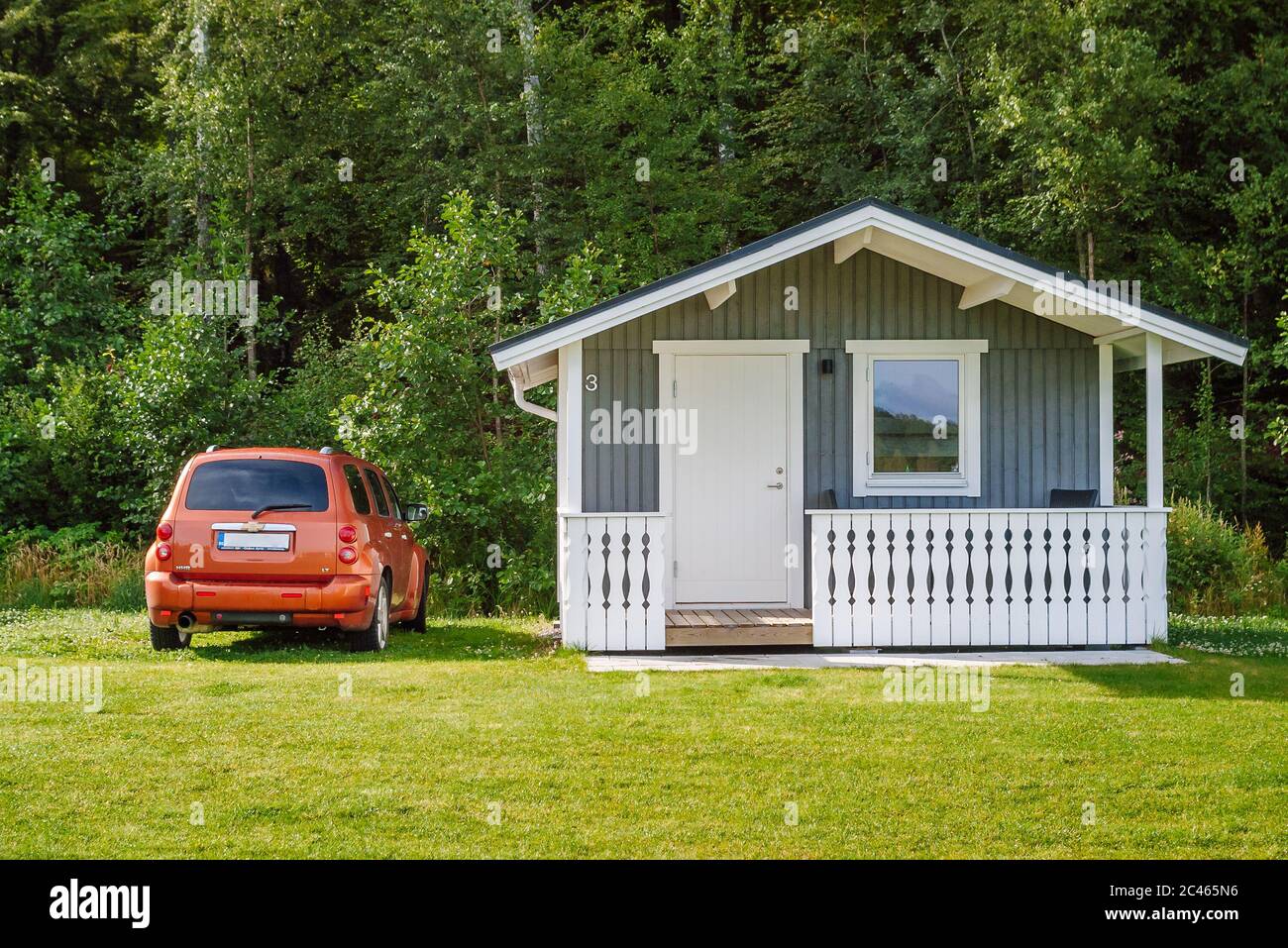 Svedese Camping, bungalow grigio in legno con recinzione bianca, parcheggiato accanto a lui un auto arancione, prato verde di fronte e fitta foresta sullo sfondo. Foto Stock