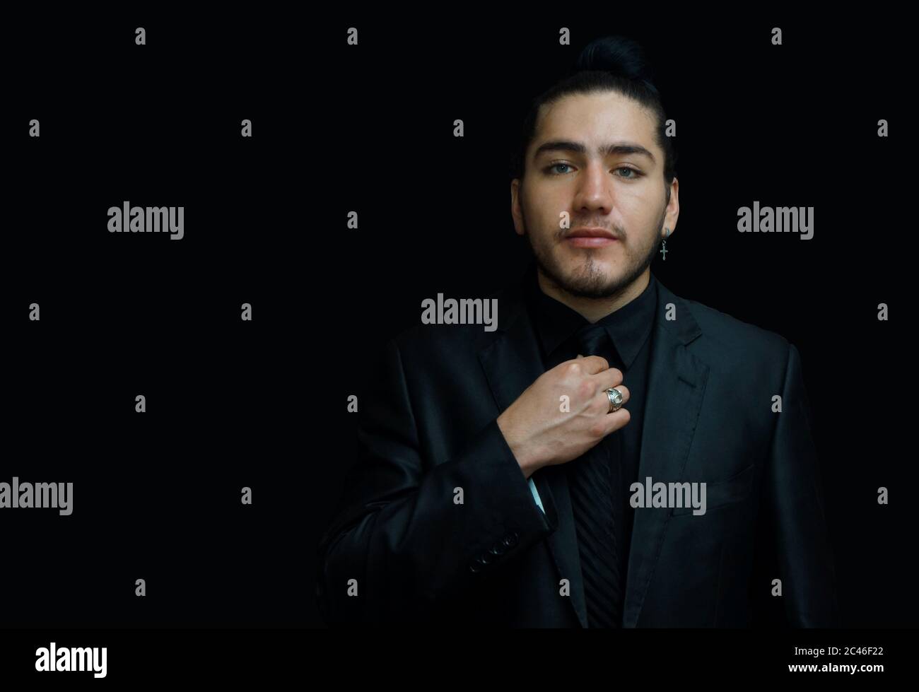 Ritratto di un giovane latino che indossa un orecchino con vestito nero, camicia  nera e cravatta nera con la mano sulla cravatta vista dalla parte anteriore  su sfondo nero Foto stock -