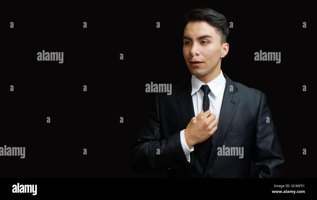 Giovane latino con abito nero, camicia bianca, cravatta nera con una mano che afferra la cravatta su sfondo nero Foto Stock