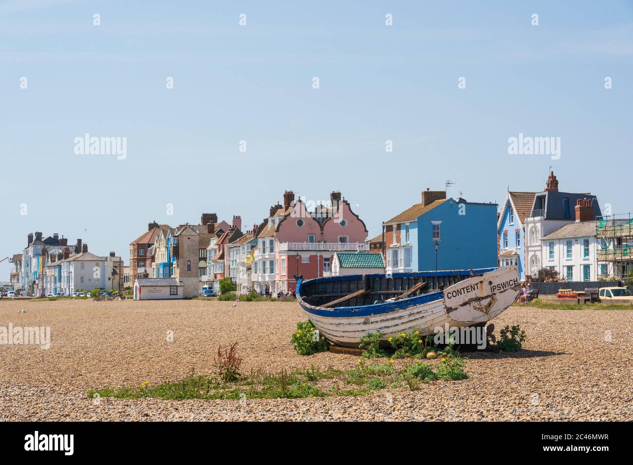 Barca da pesca abbandonata sulla spiaggia con edifici color pastello sullo sfondo in una giornata di sole con cielo blu. Aldeburgh, Suffolk. REGNO UNITO Foto Stock