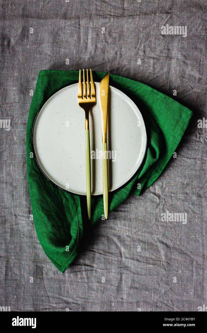 Tavolo con vista dall'alto, tela grigia da tavolo, tovagliolo di lino verde, un piatto bianco e un'elegante forchetta e coltello dorati Foto Stock
