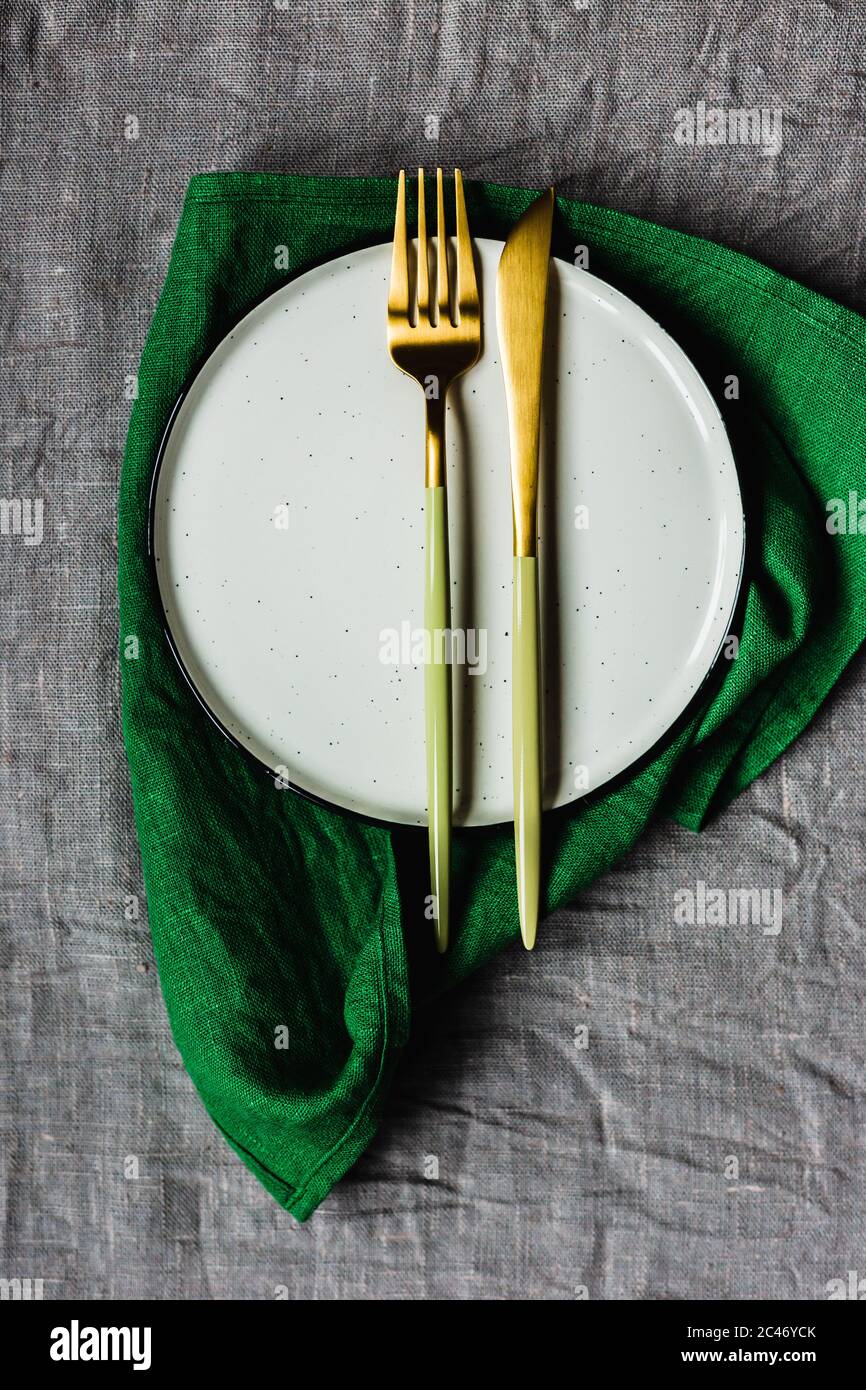 Tavolo con vista dall'alto, tela grigia da tavolo, tovagliolo di lino verde, un piatto bianco e un'elegante forchetta e coltello dorati Foto Stock