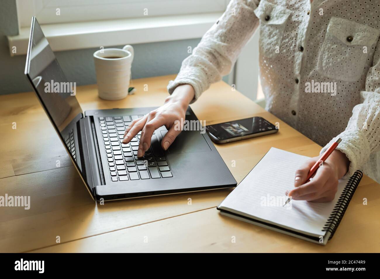 Le mani della donna che usano il computer portatile sulla scrivania all'interno della casa. Lavoro remoto. Freelance. Una tazza di tè, smartphone, notebook Foto Stock