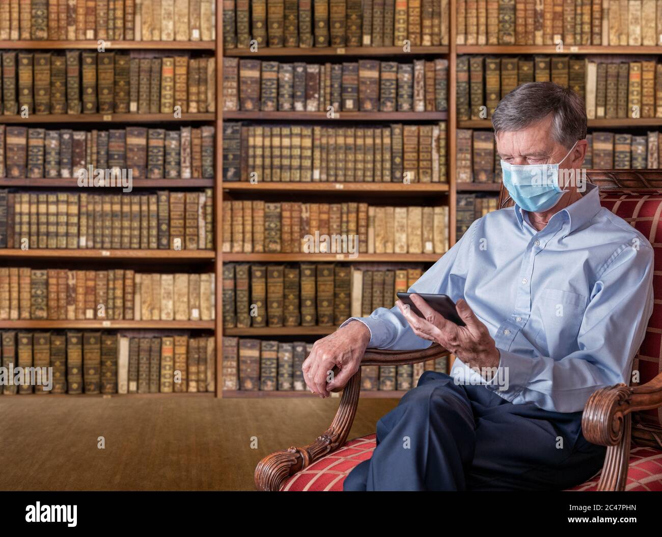 Uomo caucasico senior con una maschera contro il coronavirus che legge una tavoletta ebook in vecchia biblioteca di libri antichi Foto Stock