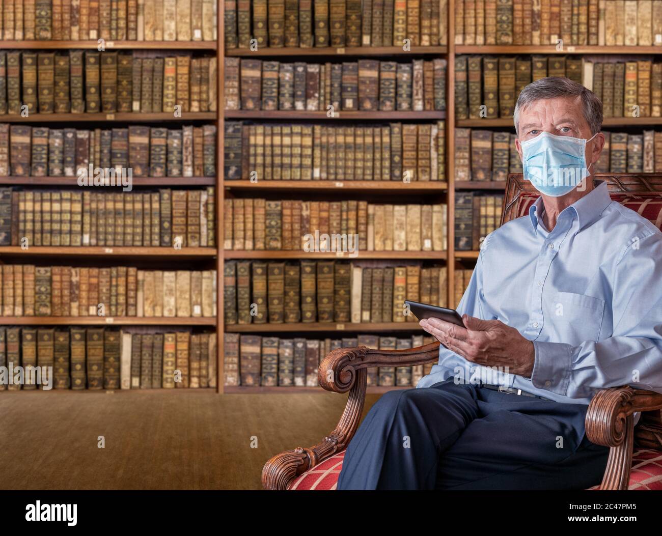 Uomo caucasico senior con una maschera contro il coronavirus che legge una tavoletta ebook in vecchia biblioteca di libri antichi Foto Stock