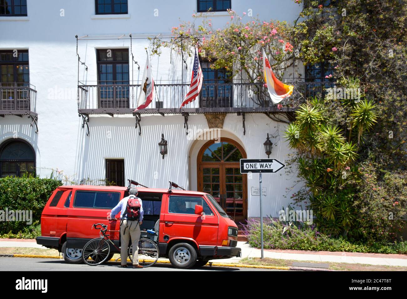 Vista posteriore di un uomo anziano che carica una bicicletta nella Volkswagen Vanagon rossa d'epoca, di fronte a un bellissimo edificio in stile spagnolo a Santa Barbara, CA, USA Foto Stock