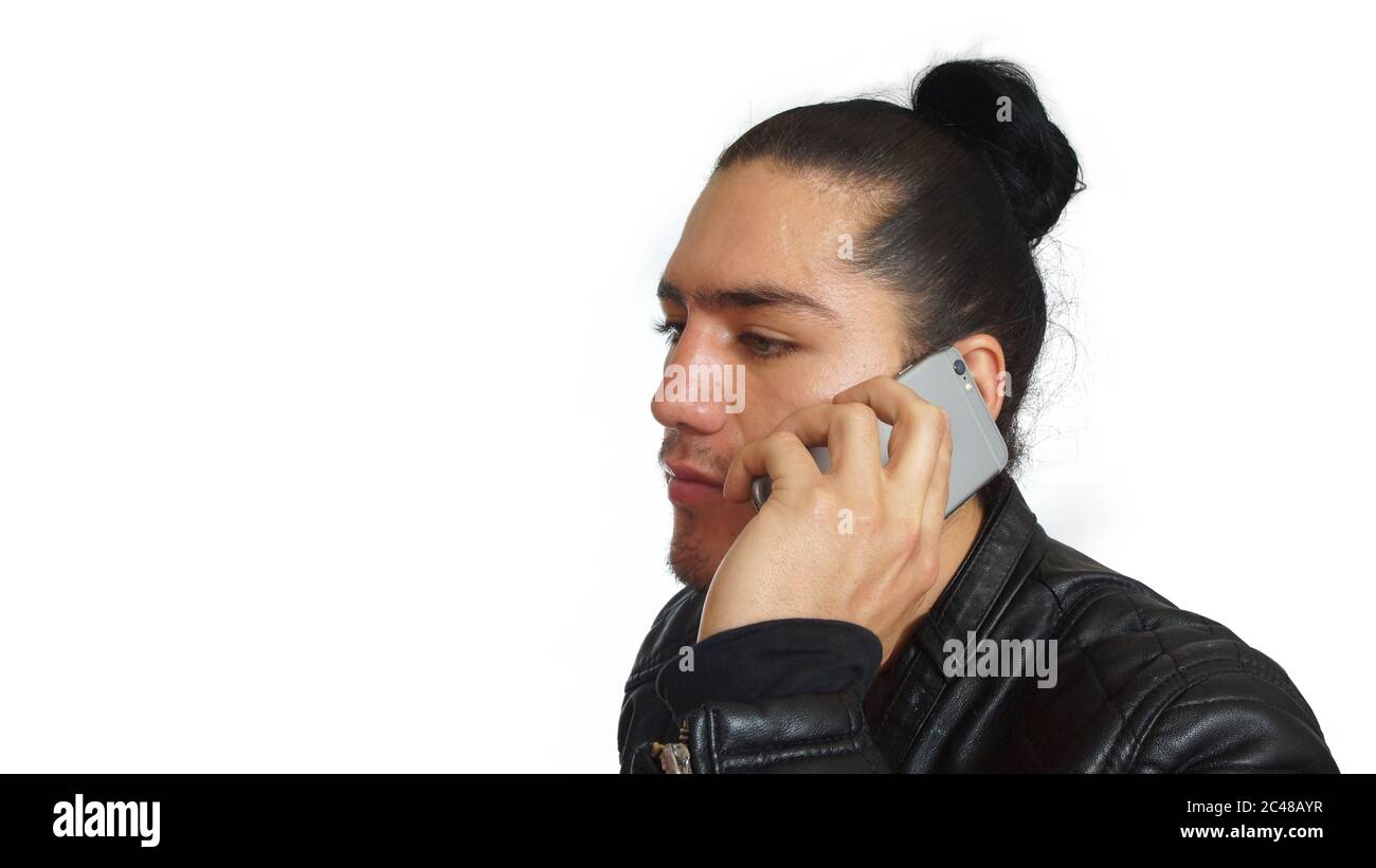Giovane uomo ispanico con capelli legati fatto in abito nero e giacca nera in pelle, con il suo cellulare in mano, visto nel profilo su sfondo bianco Foto Stock