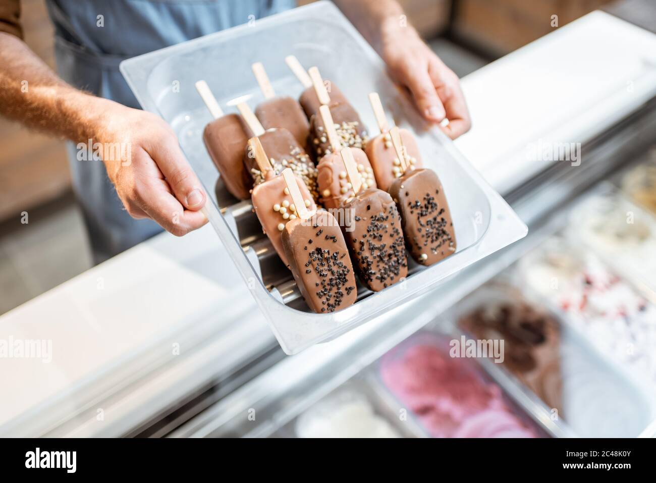 Venditore che tiene un vassoio con gelato al cioccolato su un bastone nel negozio, primo piano su gelato su uno sfondo blu grembiule Foto Stock