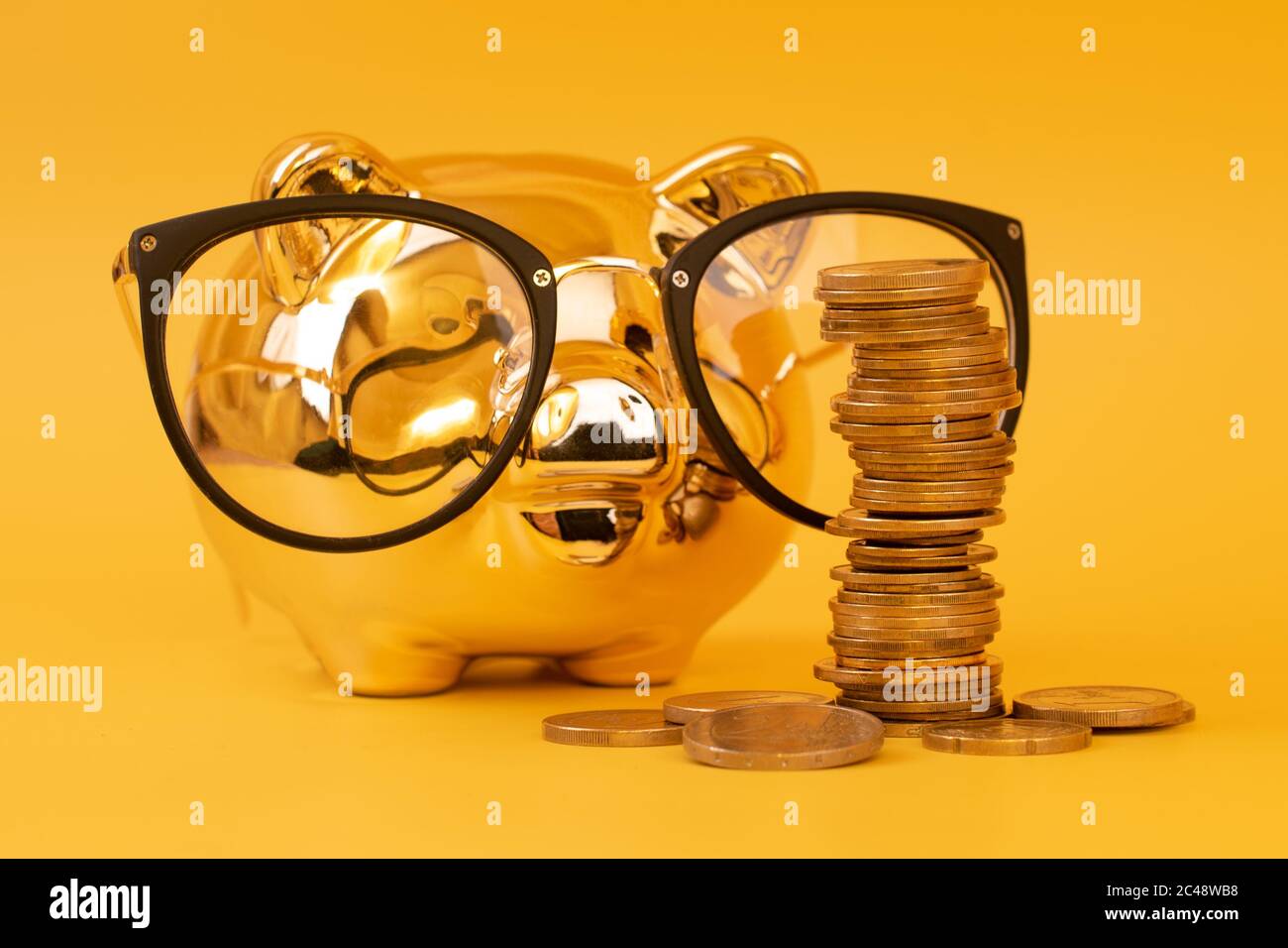 Golden piggy bank indossare occhiali con torri di denaro. Pila di monete in euro vicino al contenitore di denaro dorato. Denaro maiale, risparmio di denaro, Moneybox, mucchio di monete Foto Stock