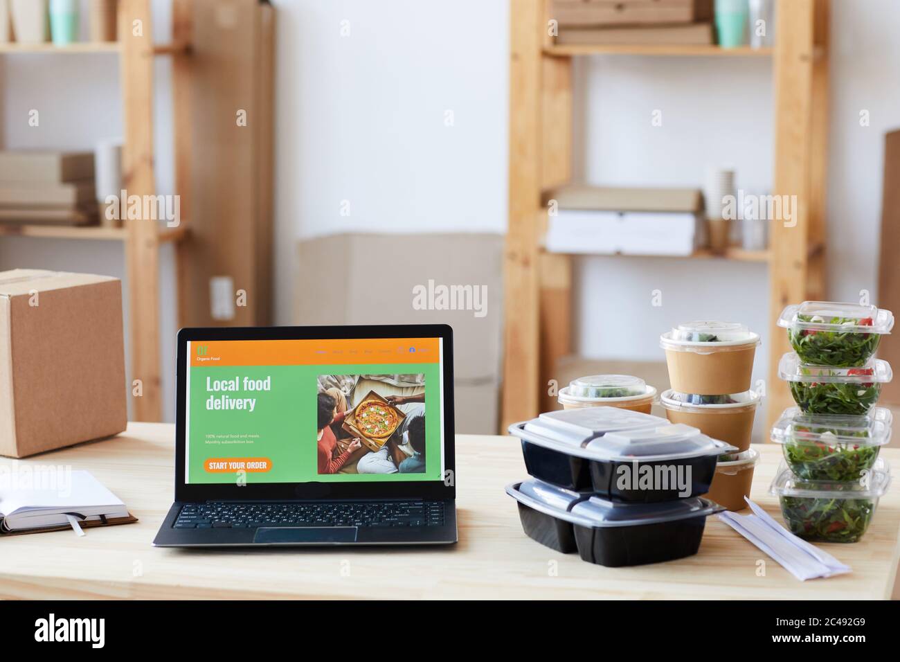 Immagine di un computer portatile con sito web del servizio di consegna di cibo e scatole di cibo sul tavolo in ufficio Foto Stock