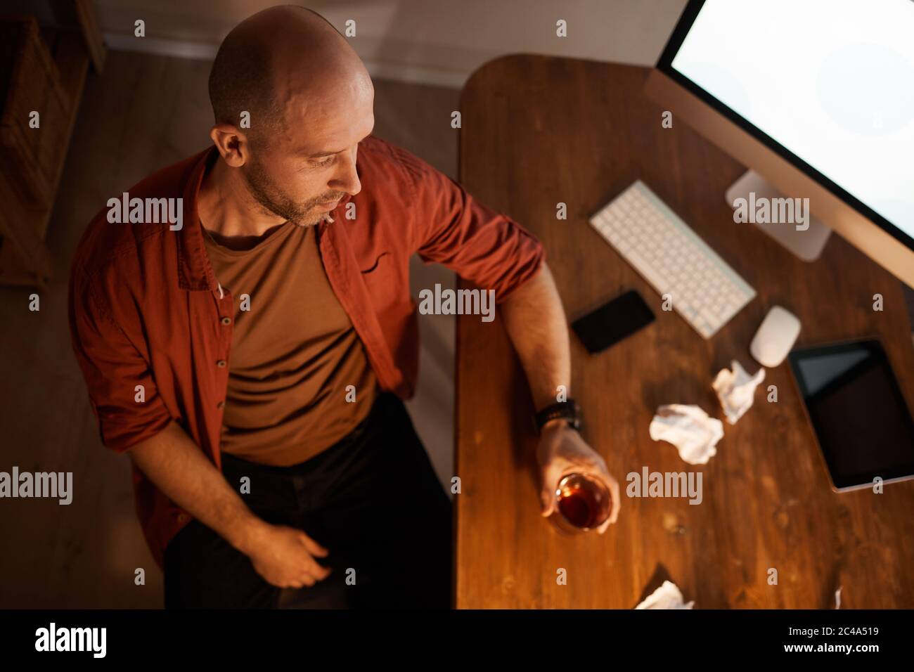 Vista ad alto angolo di un uomo maturo che beve alcol dopo una giornata di lavoro difficile, seduto sul suo posto di lavoro davanti al computer Foto Stock