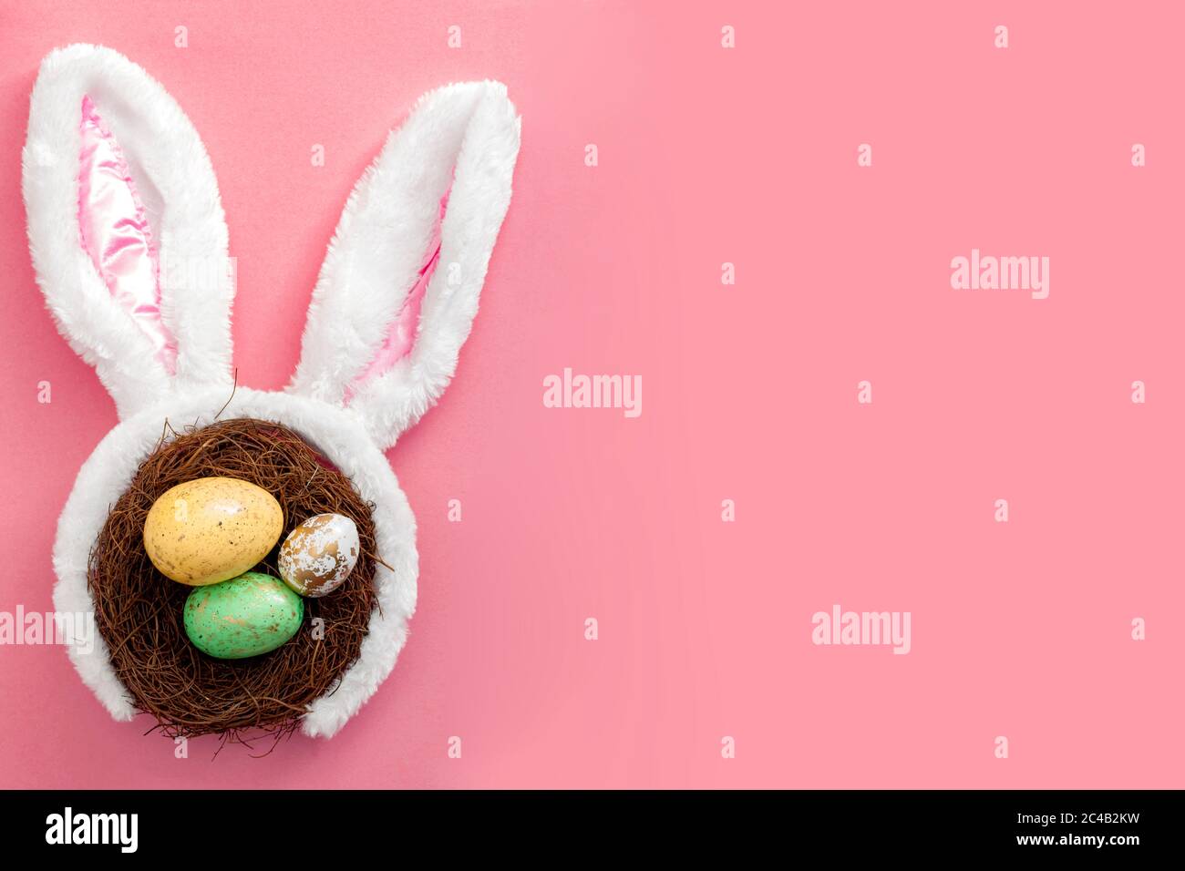 Buon concetto di memoria di Pasqua e della primavera con adorabili orecchie di coniglio su un nido di uccello con uova colorate su uno sfondo rosa luminoso con spazio di copia Foto Stock