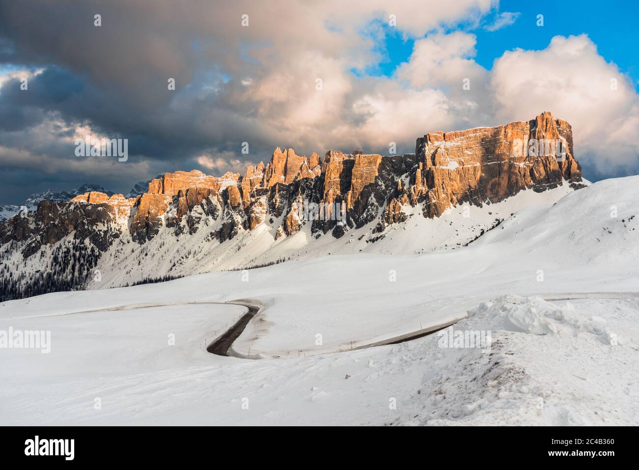 Vista dall'alto di una tortuosa strada di passaggio di montagna attraverso prati innevati e sotto torreggianti cime rocciose nelle Alpi europee al tramonto in inverno Foto Stock