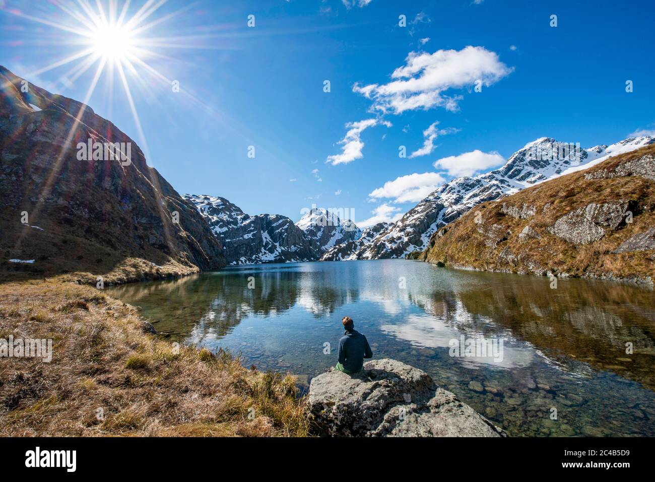 Giovane uomo seduto su una roccia, montagne riflesse nel lago, Lago Harris, Conical Hill, Routeburn Track, Mount aspirating National Park, Westland Foto Stock