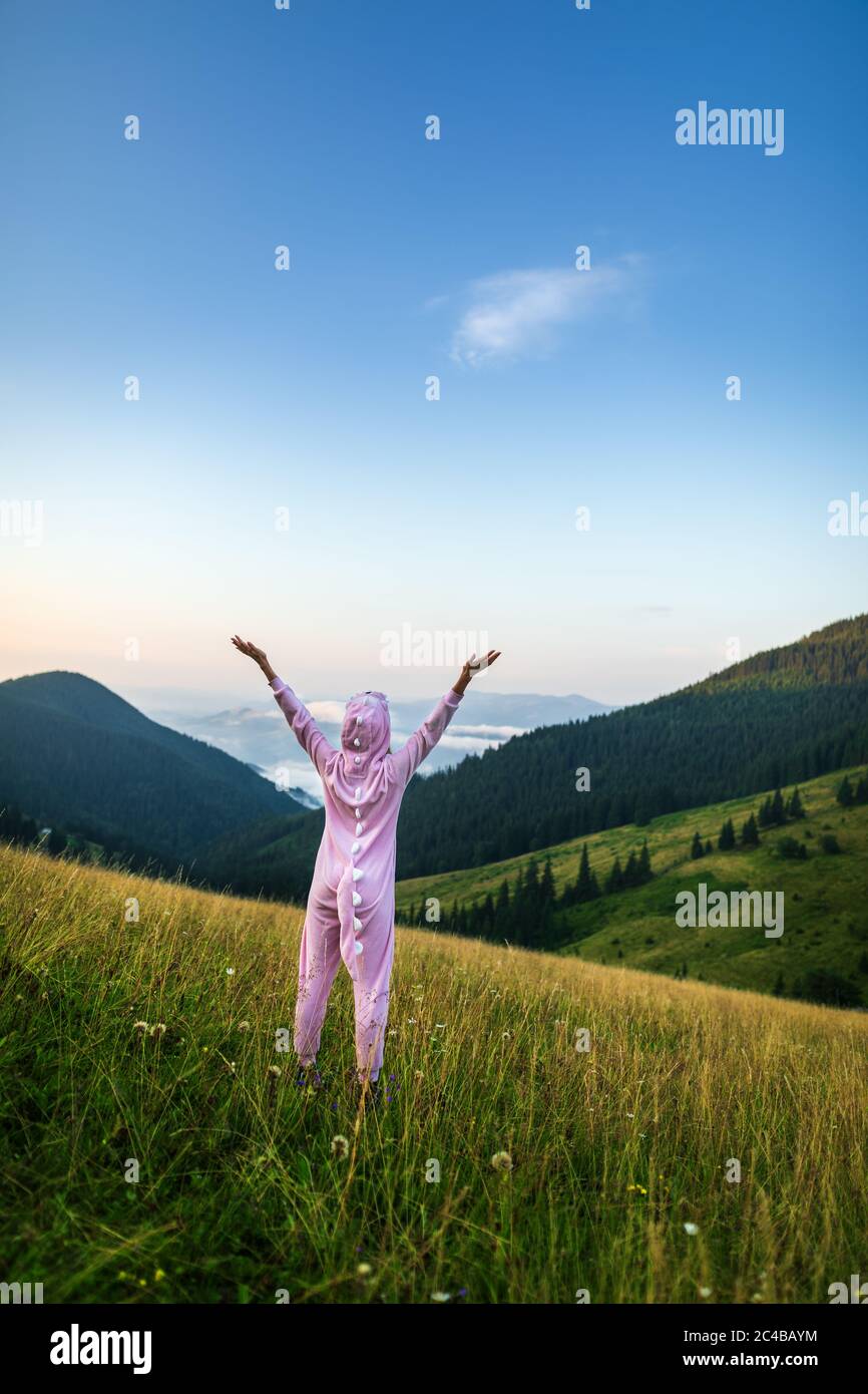 Positivo giovane donna escursionista in rosa Dragon Pajamas braccia aperte al picco di montagna. Donna sottile in tuta da notte rosa. Viaggio divertente, costume da Cosplay animale Foto Stock