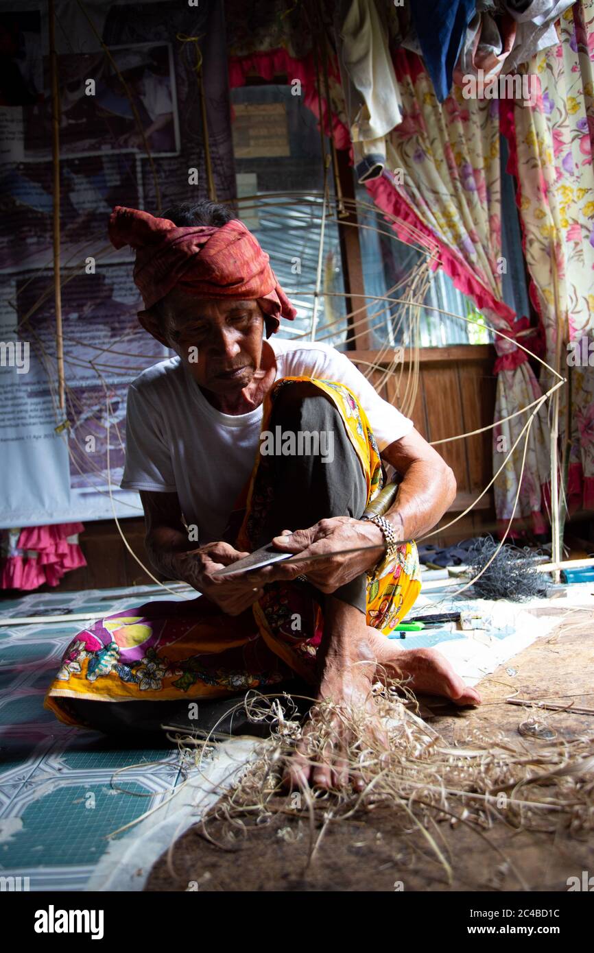 Kite maker malese che lavora su un aquilone nel suo laboratorio in condizioni di scarsa illuminazione. Foto Stock