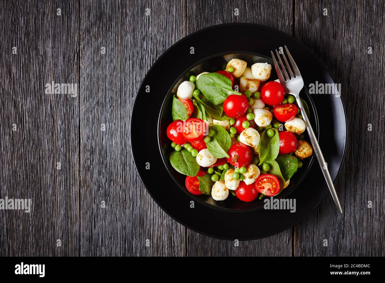 Vista dall'alto su insalata di caprese con spinaci, pomodori ciliegini, mini mozzarella, aceto balsamico e salsa all'olio d'oliva su un piatto nero, via Foto Stock