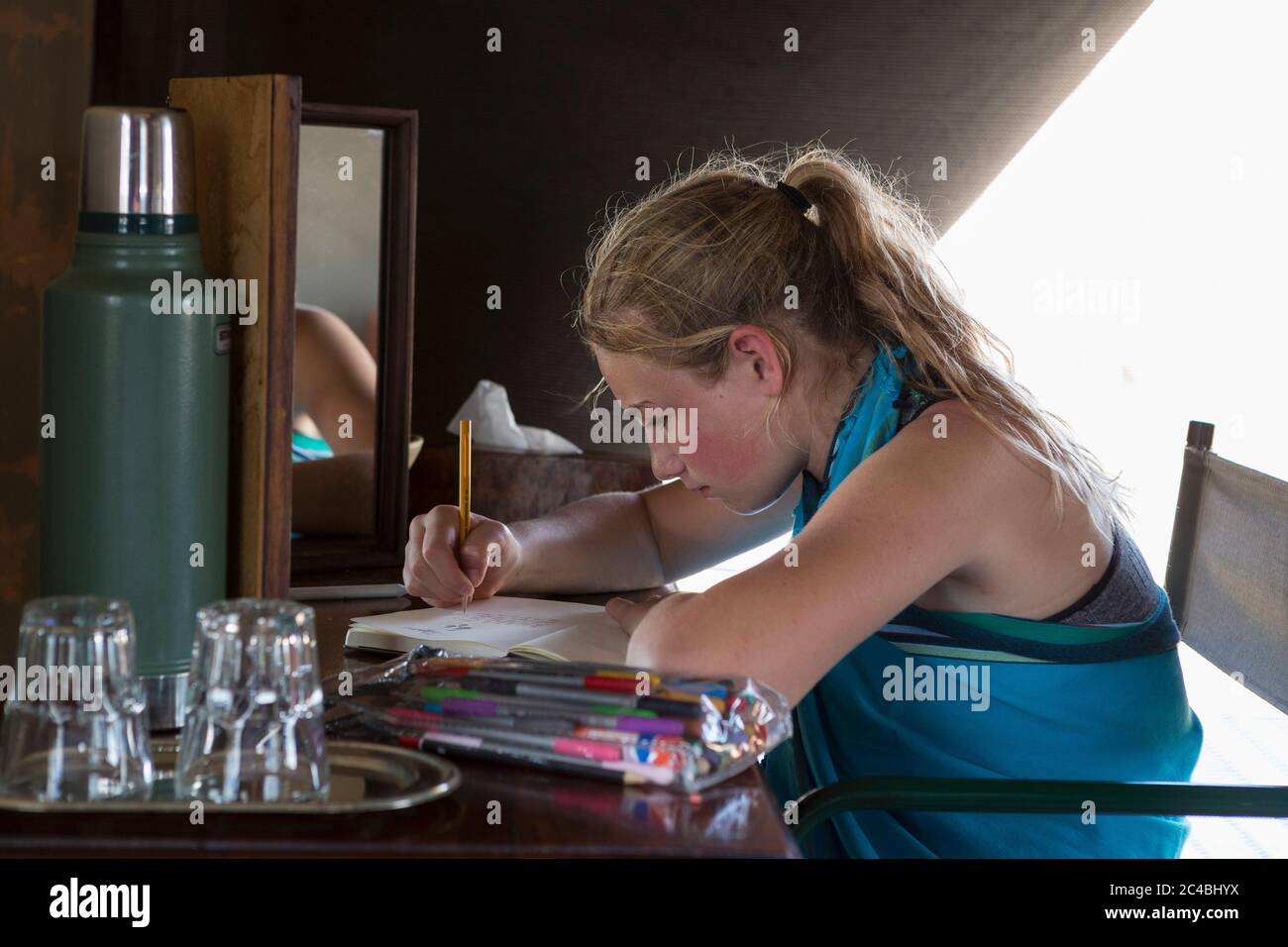 Una ragazza di dodici anni seduta su una scrivania in una tenda in un campo di riserva naturale, disegnando. Foto Stock