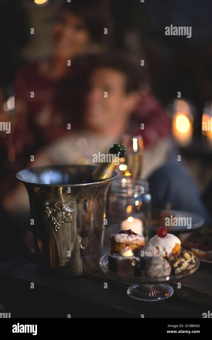 Primo piano di bottiglia di champagne in metallo di vino refrigeratore, stativo per torta di vetro con selezione di torte, persona in background. Foto Stock Primo piano di bottiglia di champagne in metallo di vino refrigeratore, stativo per torta di vetro con selezione di torte, persona in background. Foto Stock
