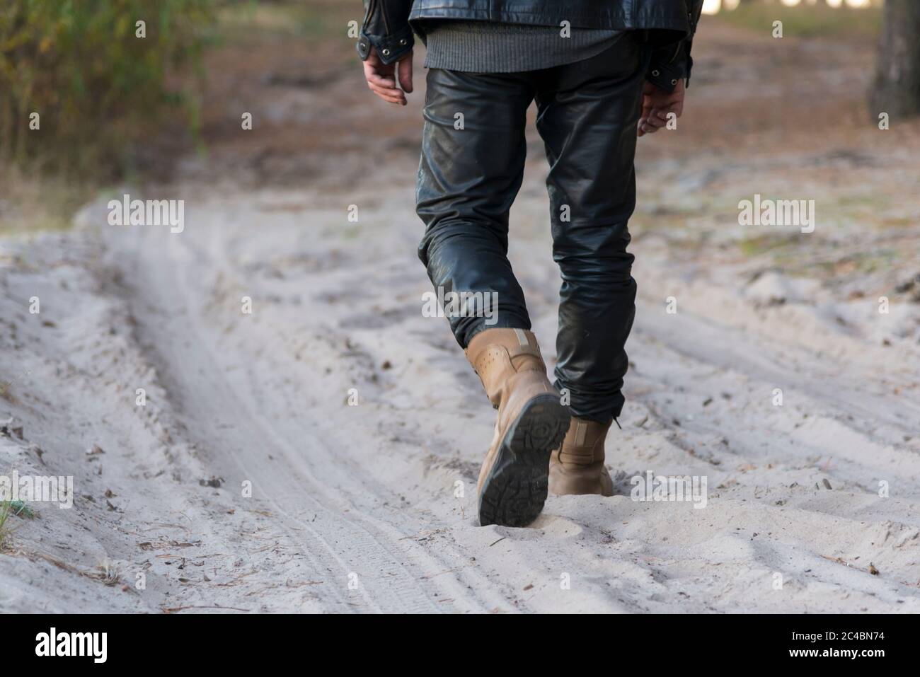 Un uomo in stivali e pantaloni in pelle sta camminando lungo una strada sabbiosa nella foresta Foto Stock