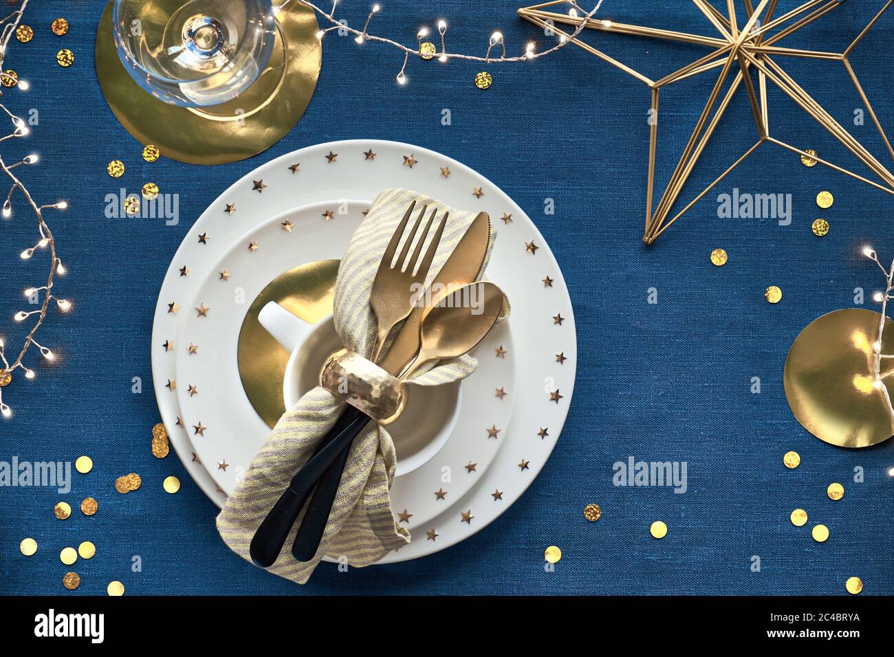 Tavolo natalizio con piatto bianco e utensili dorati e decorazioni dorate. Disposizione piatta, vista dall'alto su classico fondo tessile di lino blu. Xmas l Foto Stock