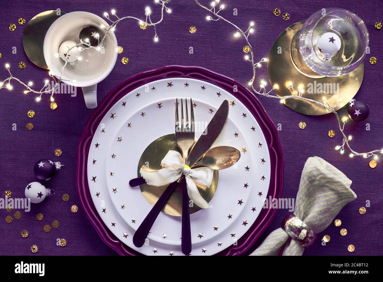 Tavolo natalizio con piatto bianco e utensili dorati e decorazioni dorate su un panno viola profondo. Disposizione piatta, vista dall'alto su biancheria viola scura Foto Stock