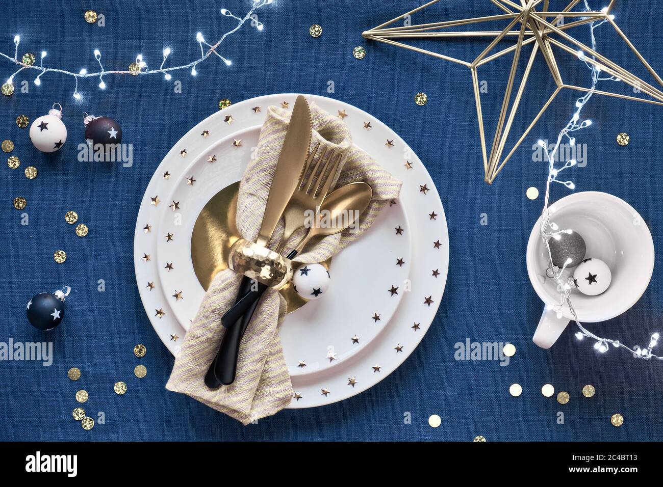 Tavolo natalizio con piatto bianco e utensili dorati e decorazioni dorate. Piatto su classico fondo tessile di lino blu. Luci di Natale garl Foto Stock