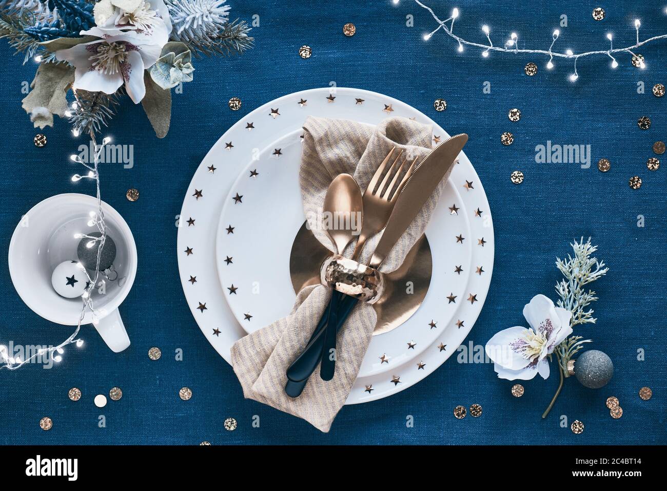 Tavolo natalizio con piatto bianco e utensili dorati e decorazioni dorate. Disposizione piatta, vista dall'alto su sfondo in tessuto di lino blu scuro. Xmas ligh Foto Stock