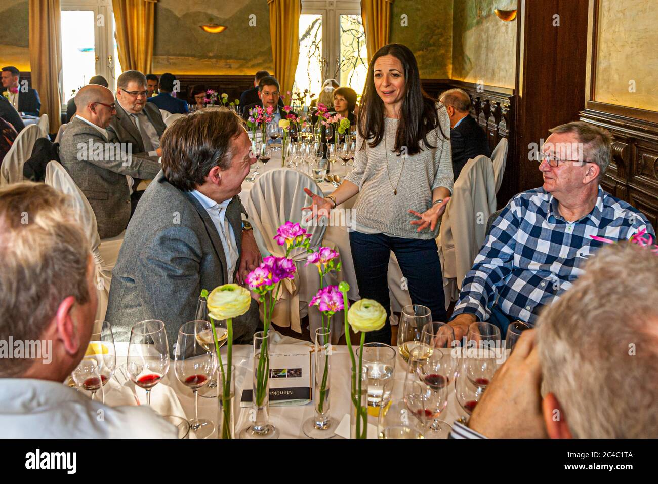 Una donna dai capelli lunghi e scuri (forse la viticoltrice Katharina Dijon della tenuta Es Fangar, Maiorca) si trova a un tavolo durante una degustazione di vini e parla agli ospiti seduti con le mani aperte nell'Hotel e Ristorante Kronenschlösschen di Hattenheim, Eltville am Rhein, Assia, Germania. Bicchieri di vino rosso e bianco, fiori in vasi sottili e menu sono appoggiati sui tavoli. Gli ospiti sono seduti a tavoli con tovaglie bianche sullo sfondo, davanti a finestre con tende dorate e affreschi. La degustazione di vini fa forse parte del pranzo di Maiorca dello chef stellato Michelin Marc Fosh (Ristorante Simply F Foto Stock