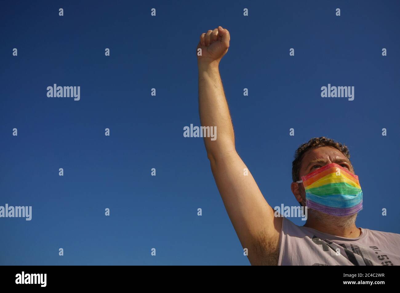 LGBTI EQUALITA Concept: Uomo con maschera da indossare a pugno con bandiera iridata lgbt nella prevenzione del coronavirus combattendo per i suoi diritti sul cielo blu Foto Stock