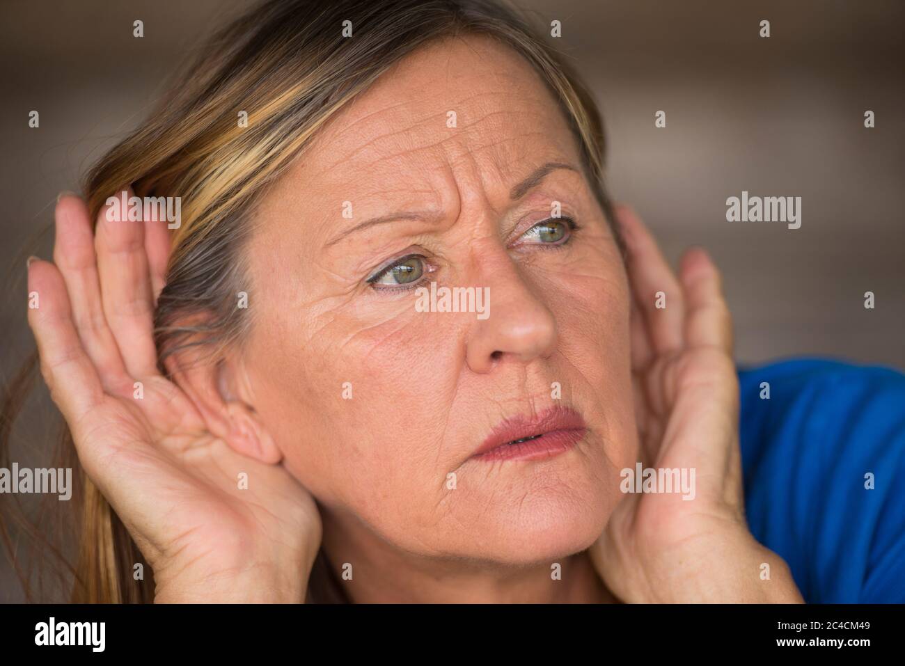 Ritratto attraente donna matura con le mani alle orecchie ascoltando con curioso, interessato e preoccupato espressione facciale a forte rumore o suono, sfocato b Foto Stock