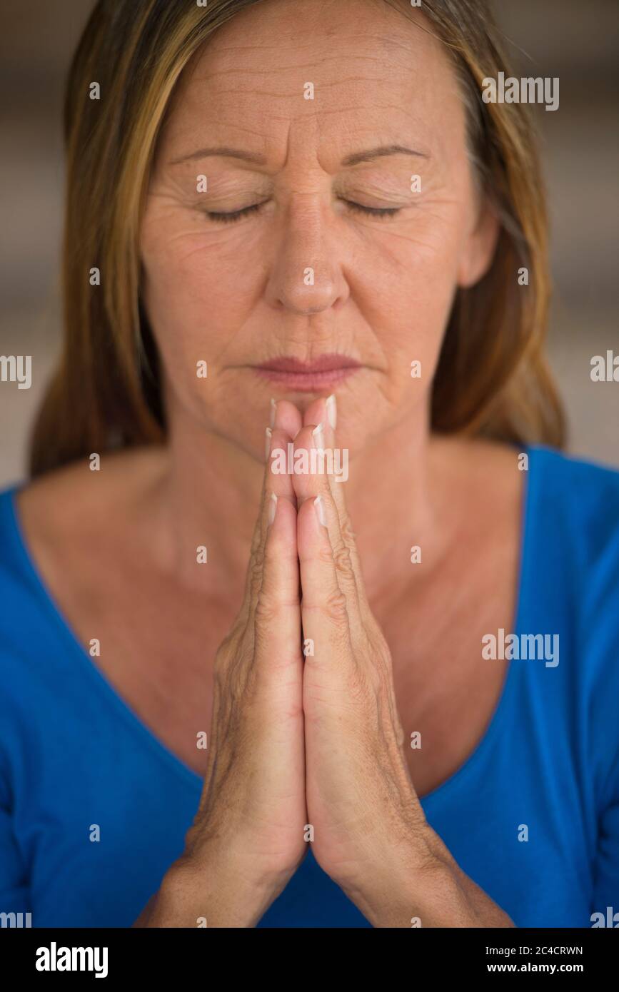 Portrtait attraente donna religiosa matura che prega con il fuoco sulle mani piegate, premuroso, pacifico, meditante, occhi chiusi, sfondo sfocato. Foto Stock