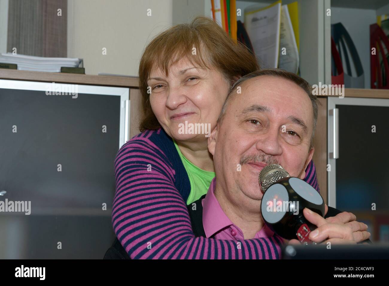 Primo piano di un ritratto indoor coppia di uomo anziano che firma con microfono e la sua ragazza che sta mettendo le braccia intorno all'uomo dalla sua schiena. Foto Stock