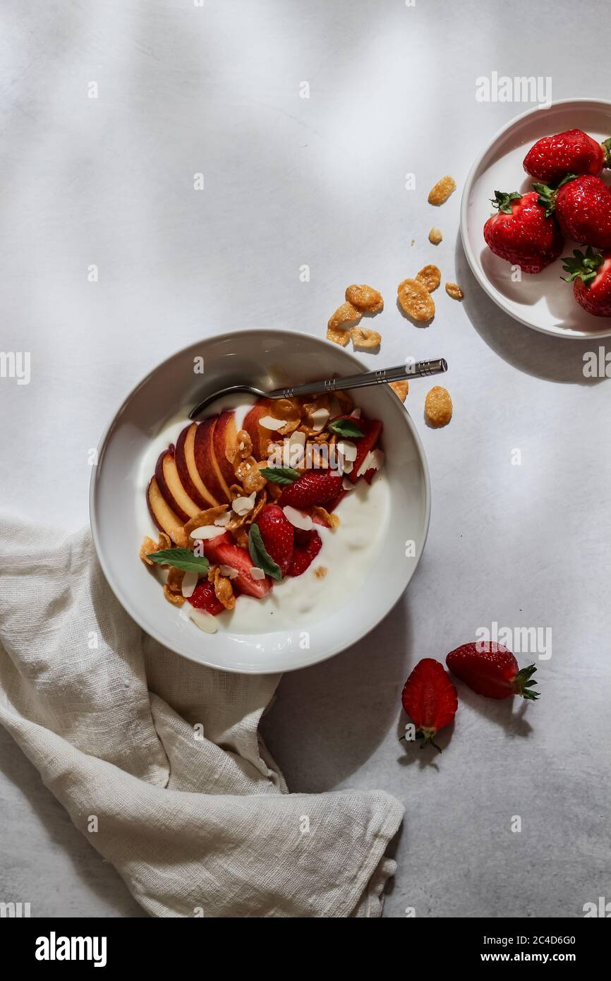 Ciotola per la colazione con yogurt, fragole, pesca a fette e fiocchi su un tavolo bianco Foto Stock