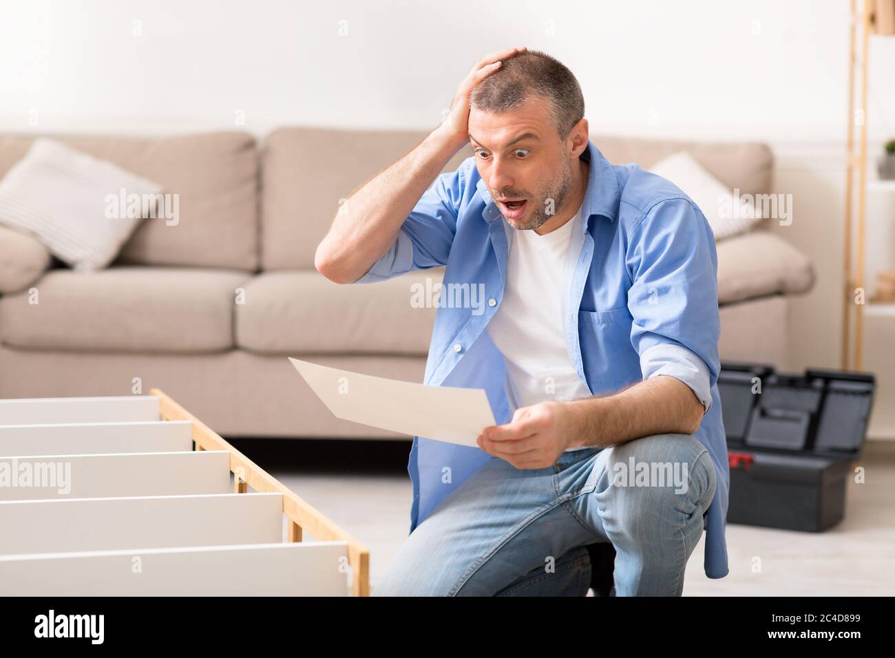 Uomo scioccato guardando le istruzioni di installazione di scaffale testa graffiante in interni Foto Stock