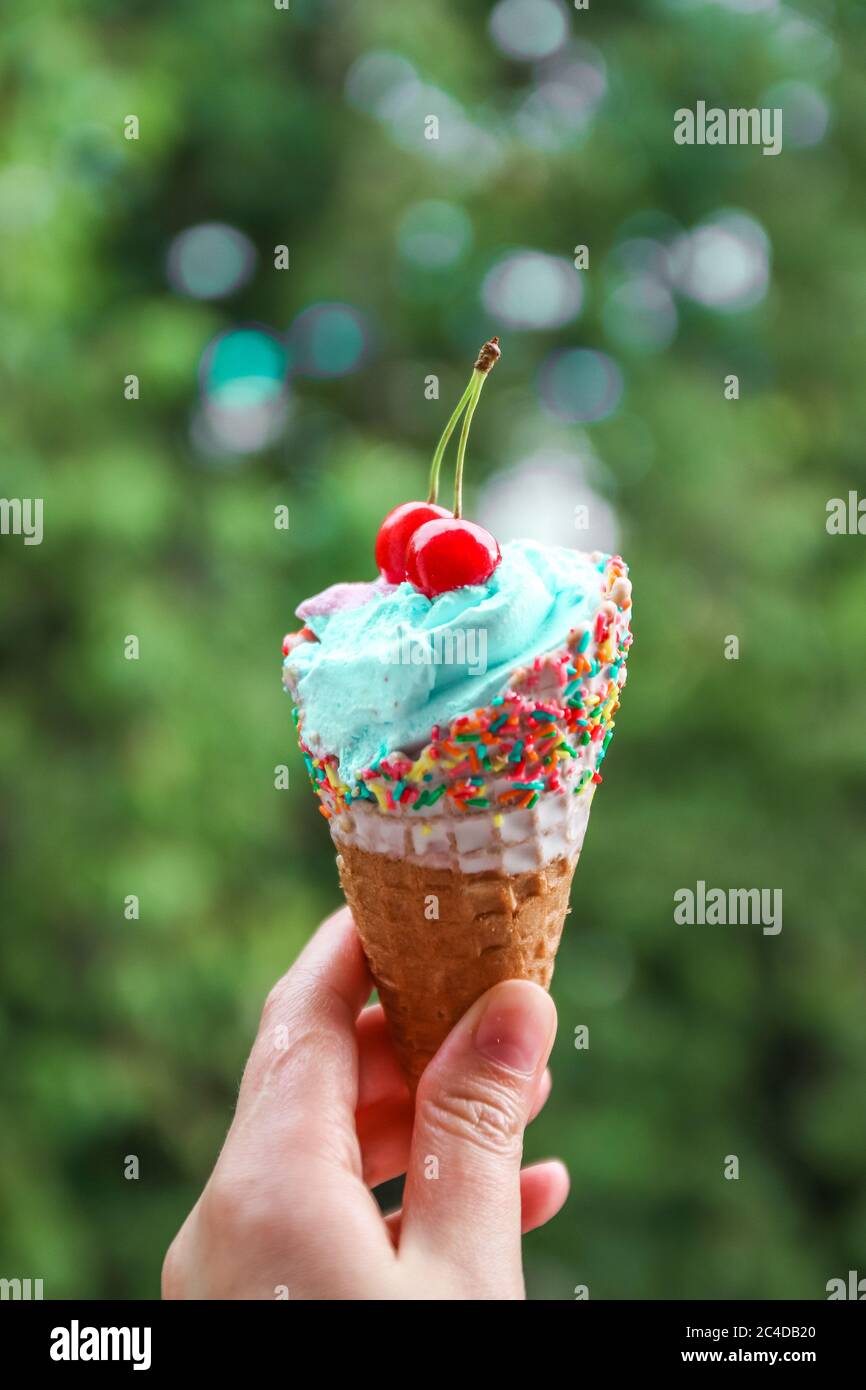 La mano femminile tiene il gelato multicolore con le ciliegie in un cono di cialda contro sfondo verde sfocato Foto Stock