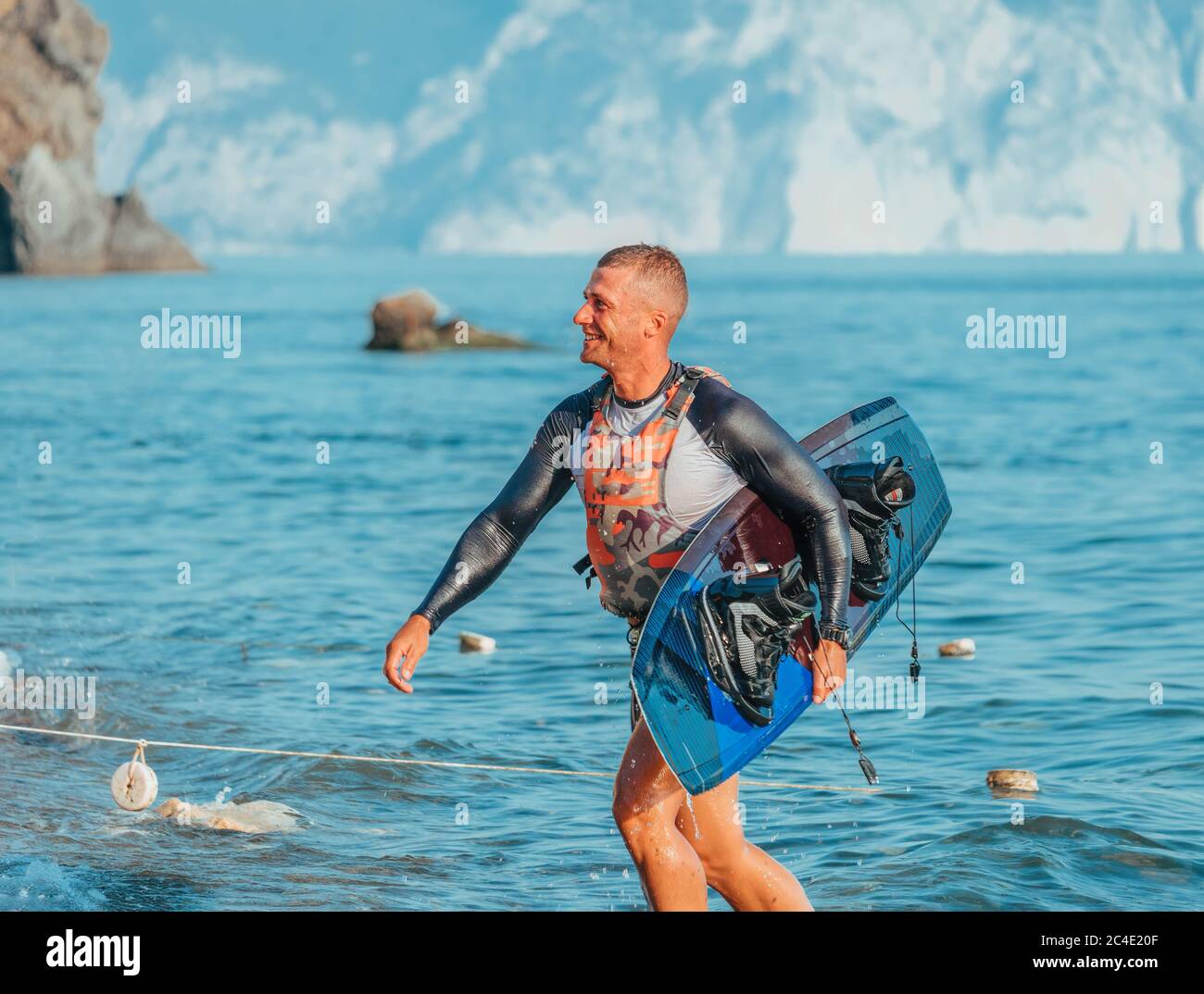 Felice uomo sorridente con un tavolo per wake surf sulla spiaggia dopo aver cavalcato al tramonto. Sci d'acqua. Un atleta maschile gioca sport acquatici con una tavola. Sveglia Foto Stock