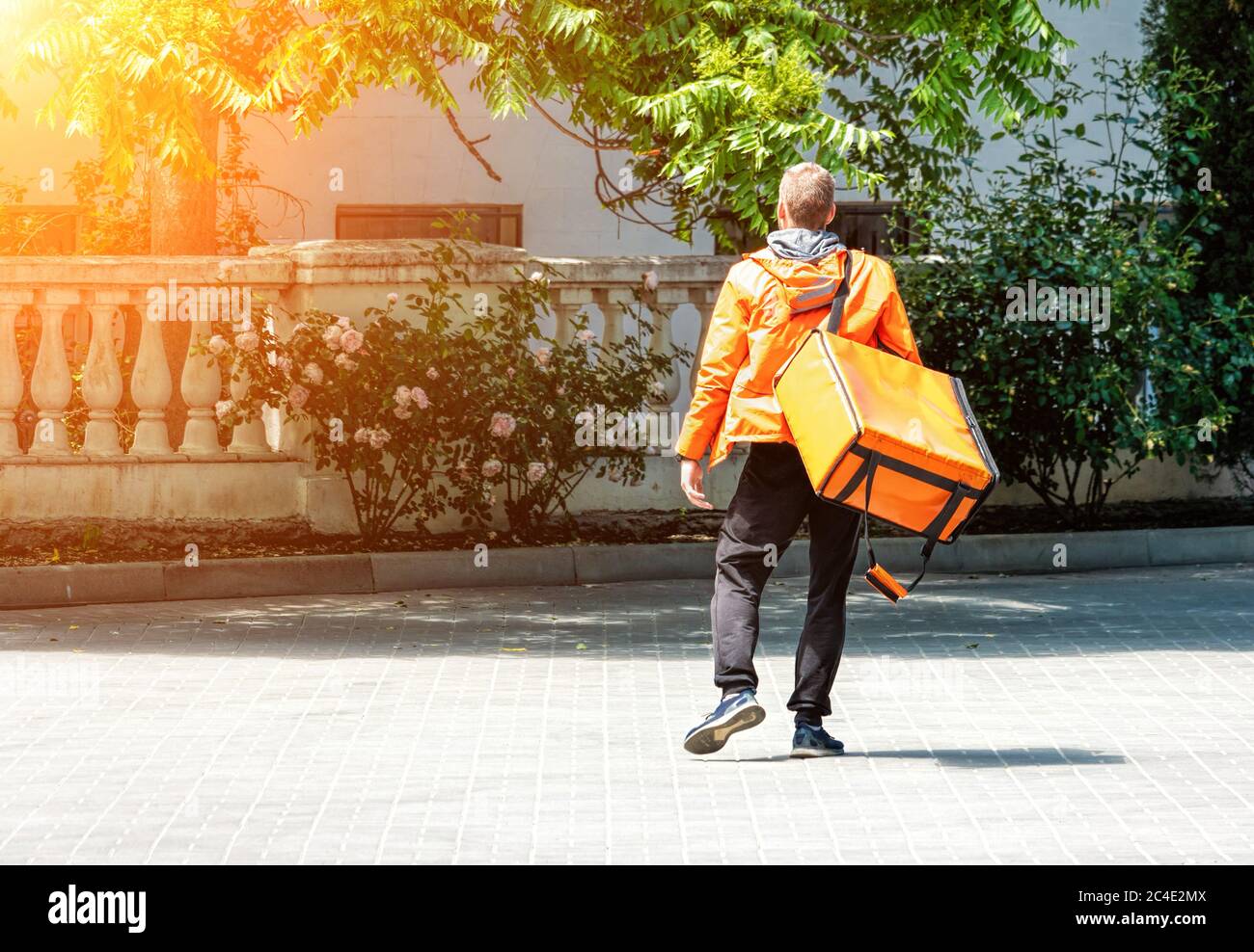 Uomo di consegna con zaino. Deliveryman caucasico isolato in uniforme arancione su sfondo parco. Servizio di consegna senza contatto durante il corso Foto Stock