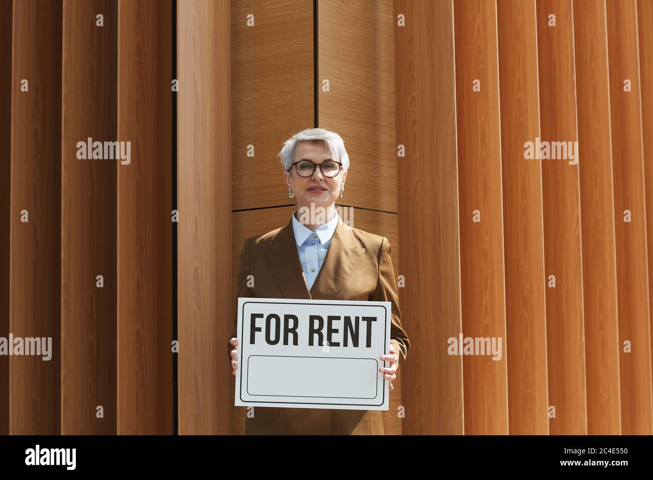 Ritratto di agente immobiliare maturo in occhiali tenendo un cartello per l'affitto e sorridendo a macchina fotografica all'aperto Foto Stock