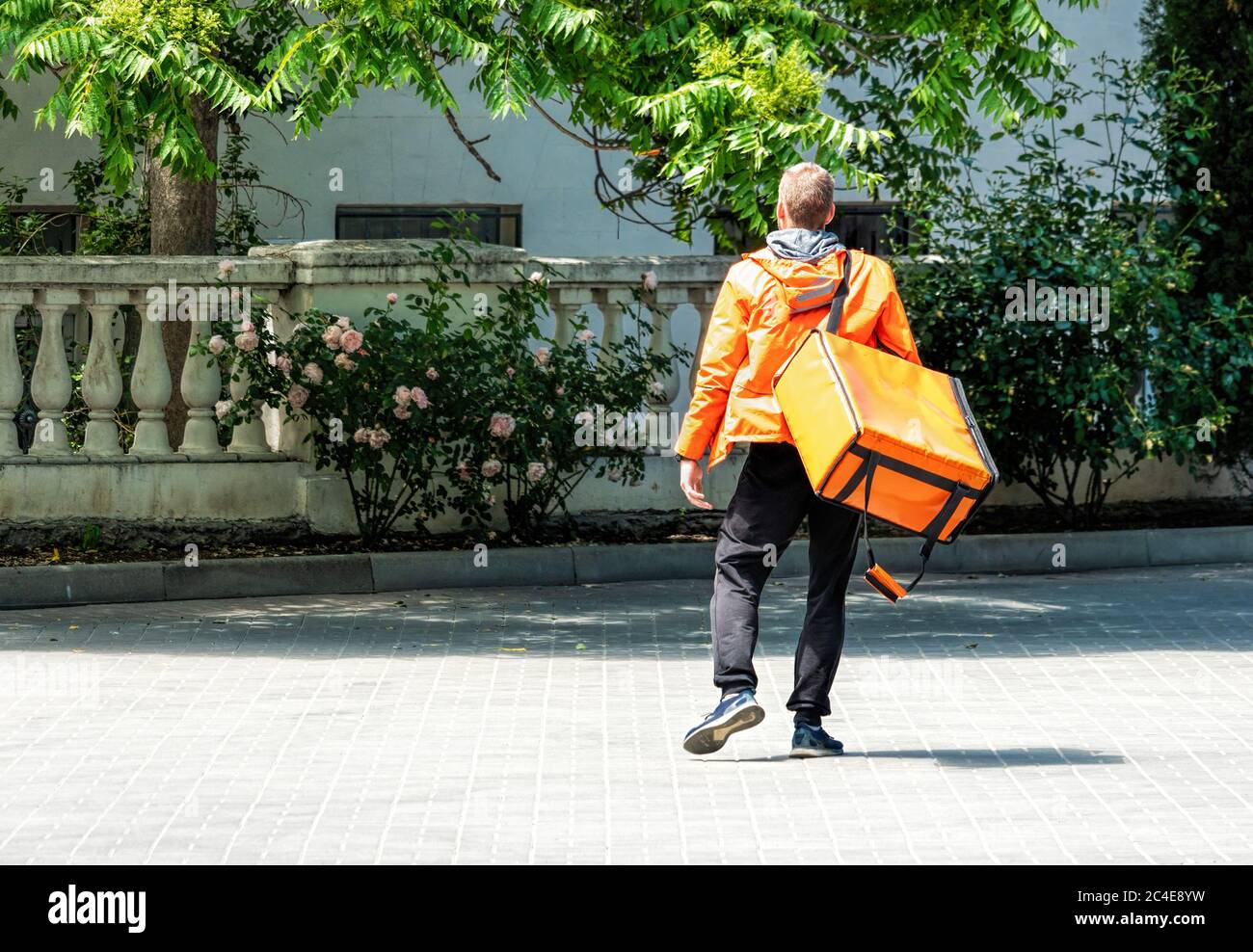 Uomo di consegna con zaino. Deliveryman caucasico isolato in uniforme arancione su sfondo parco. Servizio di consegna senza contatto durante il corso Foto Stock