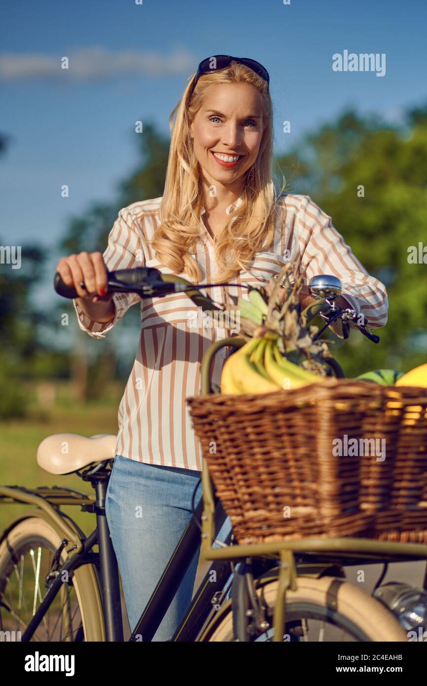 Donna che usa la sua bicicletta per acquistare prodotti freschi in piedi su una strada rurale nel caldo bagliore del sole sorridendo felice alla macchina fotografica come lei tiene la sua moto Foto Stock